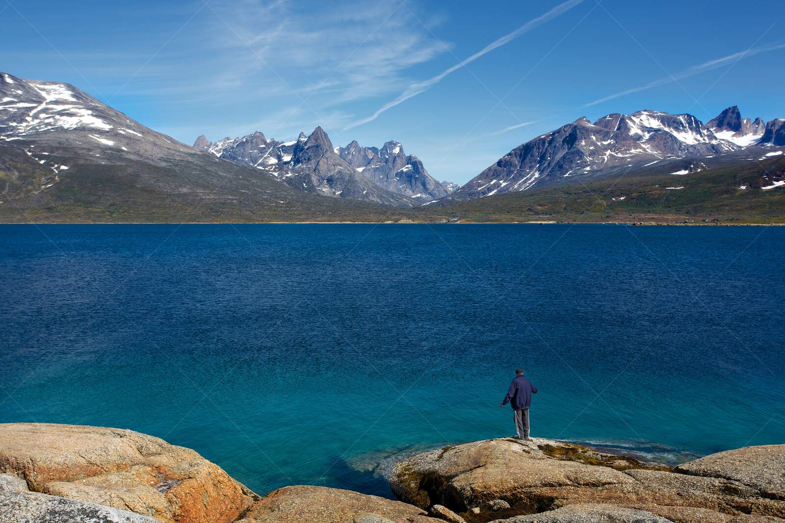 Man standing on rocky shore by blue lake with snowy mountains